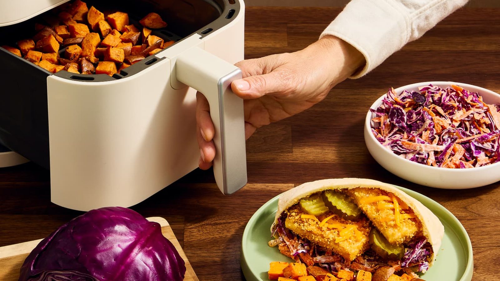 An air fryer of sweet potatoes beside a loaded pita and a bowl of red cabbage slaw