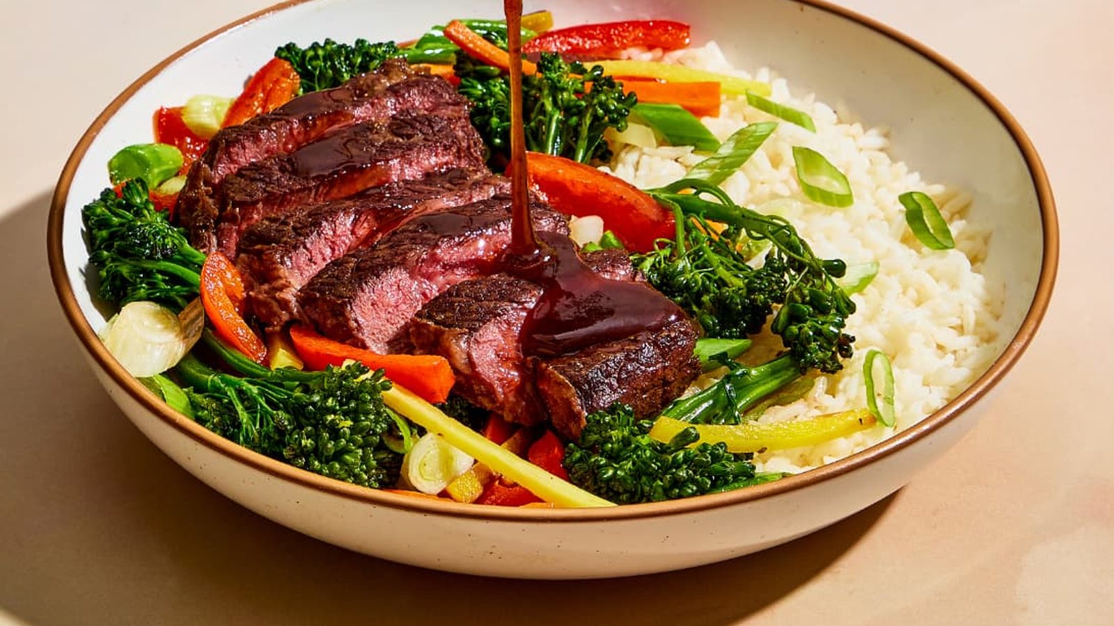 Sliced steak with broccolini and rice in a bowl, sauce being poured over the top