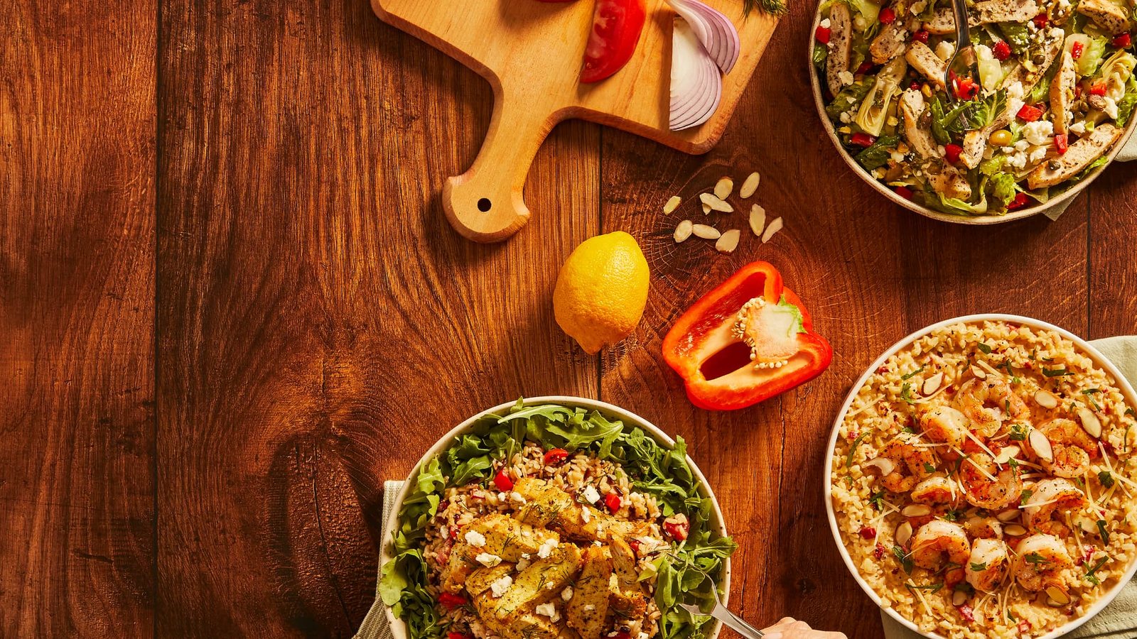 Three Green Chef bowls on a wooden table beside a cutting board of tomato, onion and herbs