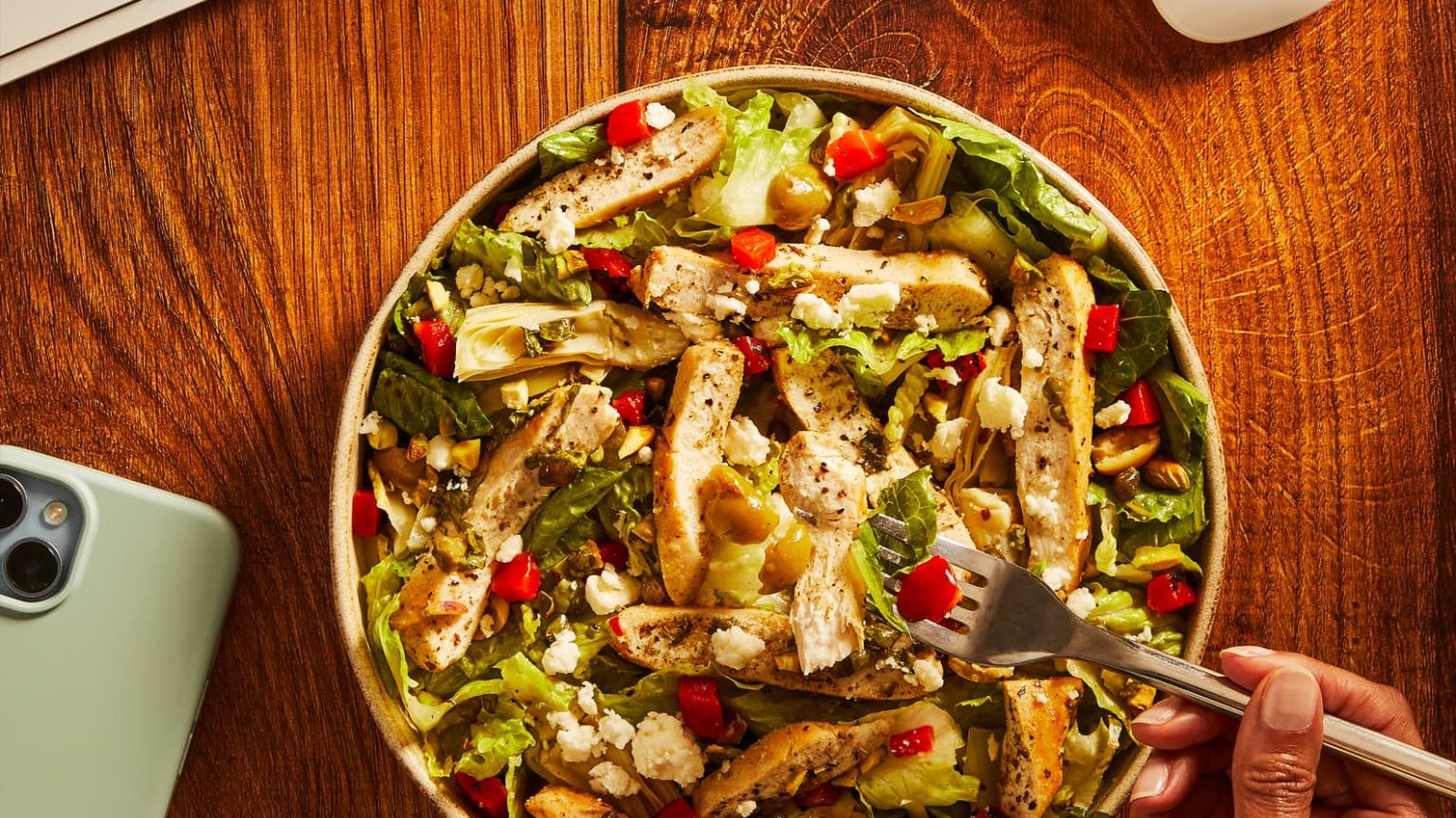 A chicken, artichoke and feta salad bowl on a desk beside a keyboard and phone